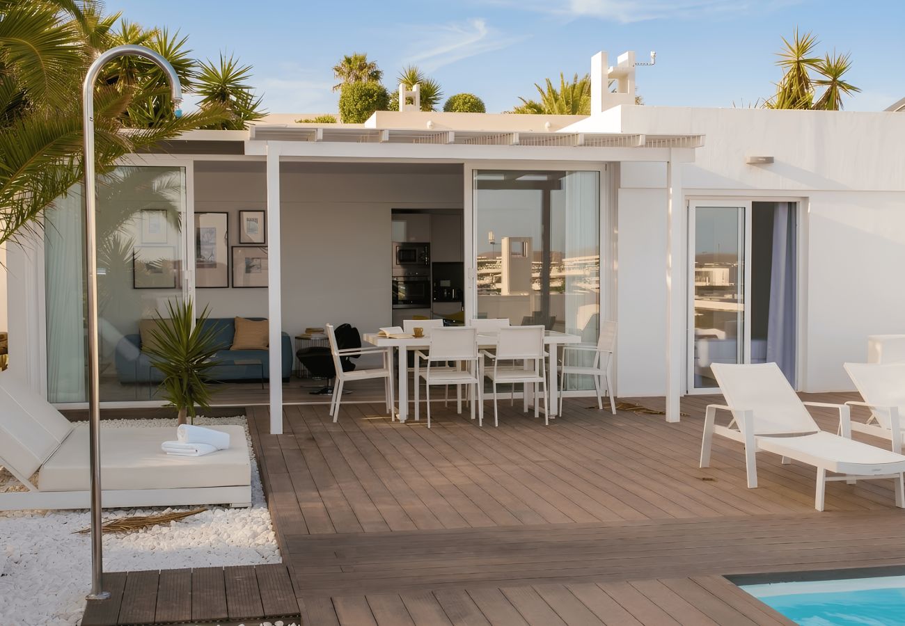 Terraza exterior con suelo de madera, ducha, tumbonas blancas y mesa bajo pérgola